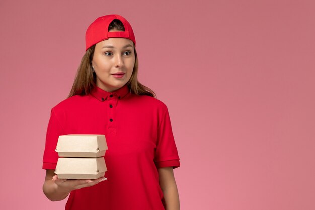 Mensajero femenino de vista frontal en uniforme rojo y capa sosteniendo paquetes de comida de entrega en la pared rosa claro, trabajo de trabajo de empresa de servicio de entrega uniforme