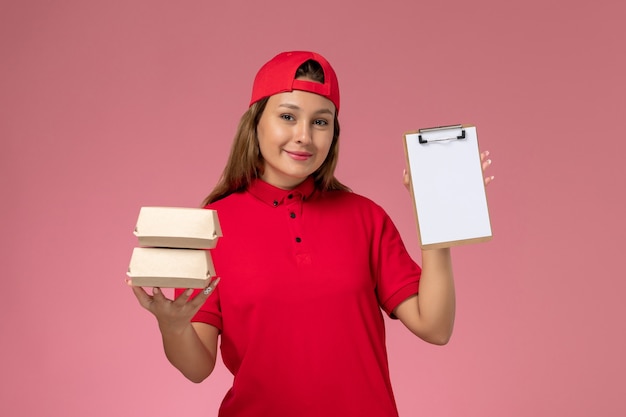 Mensajero femenino de vista frontal en uniforme rojo y capa sosteniendo un pequeño bloc de notas de paquetes de comida de entrega en la pared rosa claro, servicio de entrega de trabajo de trabajador uniforme