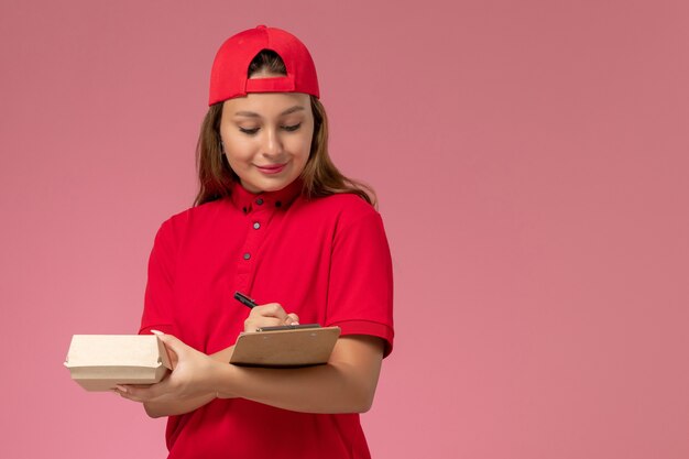 Mensajero femenino de vista frontal en uniforme rojo y capa sosteniendo un pequeño paquete de comida de entrega y un bloc de notas escrito en la pared rosa, servicio de entrega uniforme de trabajo