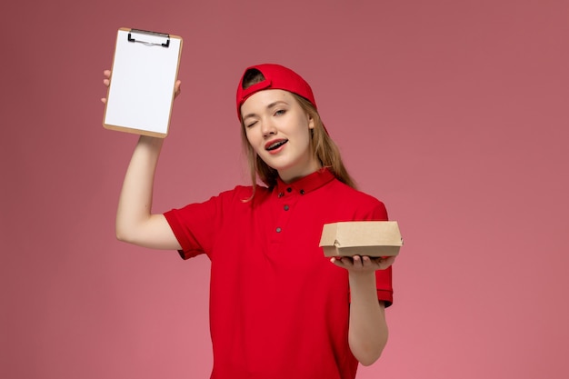 Mensajero femenino de vista frontal con uniforme rojo y capa sosteniendo un pequeño paquete de comida de entrega con bloc de notas guiñando un ojo en la pared rosa, uniforme de trabajo de la empresa de servicios de entrega