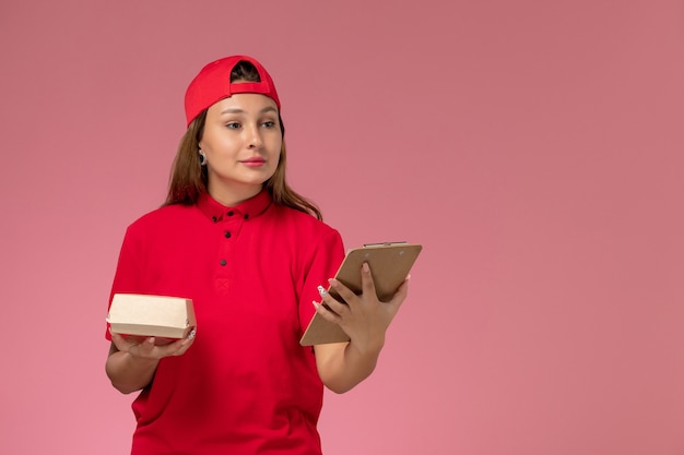 Mensajero femenino de vista frontal en uniforme rojo y capa sosteniendo un pequeño paquete de comida de entrega y un bloc de notas en la pared rosa, empresa de servicio de entrega de uniforme de trabajo