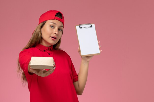 Mensajero femenino de vista frontal en uniforme rojo y capa sosteniendo un pequeño paquete de comida de entrega y un bloc de notas en la pared rosa, empresa de servicio de entrega de uniforme de trabajo