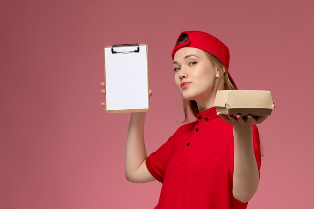 Mensajero femenino de vista frontal en uniforme rojo y capa sosteniendo un pequeño paquete de comida de entrega con bloc de notas en la pared rosa, servicio de entrega uniforme trabajo trabajador
