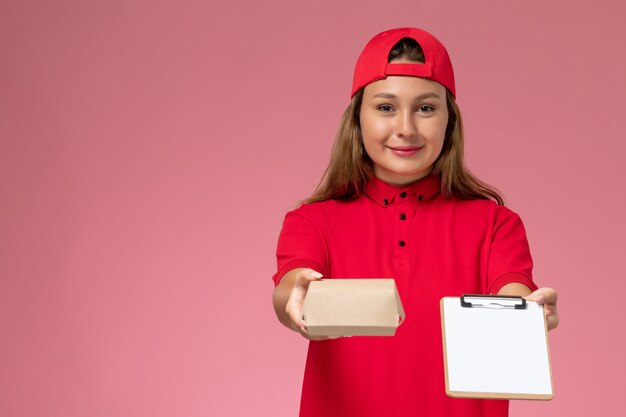 Mensajero femenino de vista frontal en uniforme rojo y capa sosteniendo un pequeño paquete de comida de entrega y un bloc de notas en la pared rosa, trabajo de servicio de entrega uniforme