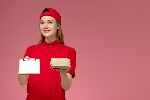 Mensajero femenino de vista frontal en uniforme rojo y capa sosteniendo un pequeño paquete de comida de entrega con bloc de notas sonriendo en la pared rosa, servicio de entrega uniforme
