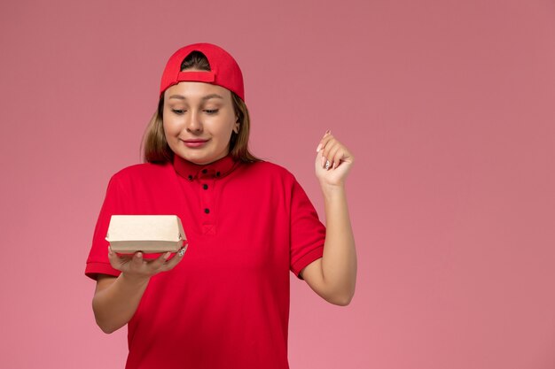 Mensajero femenino de vista frontal en uniforme rojo y capa sosteniendo un pequeño paquete de comida de entrega en una empresa de servicio de entrega uniforme de escritorio rosa claro