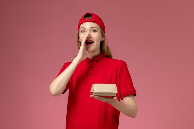 Mensajero femenino de vista frontal con uniforme rojo y capa sosteniendo un pequeño paquete de comida de entrega llamando en la pared rosa, trabajo uniforme de la empresa de servicios de entrega