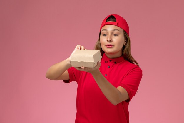 Mensajero femenino de vista frontal en uniforme rojo y capa sosteniendo un pequeño paquete de comida de entrega en la pared de color rosa claro, trabajador de la empresa de servicios de entrega uniforme