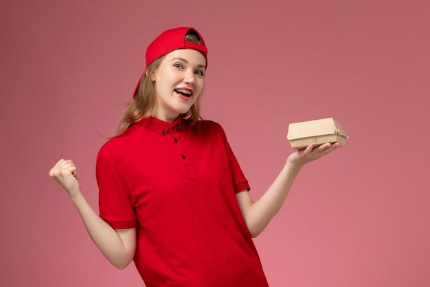 Mensajero femenino de vista frontal en uniforme rojo y capa sosteniendo un pequeño paquete de comida de entrega en una pared rosa claro, trabajador de trabajo uniforme de la empresa de servicios de entrega