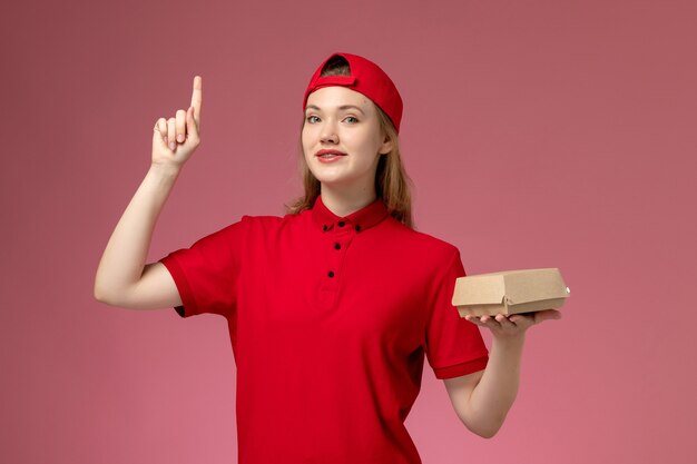 Mensajero femenino de vista frontal en uniforme rojo y capa sosteniendo un pequeño paquete de comida de entrega en la pared rosa claro, trabajo uniforme de la empresa de servicios de entrega