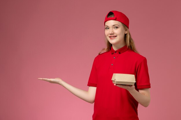 Mensajero femenino de vista frontal en uniforme rojo y capa sosteniendo un pequeño paquete de comida de entrega en la pared rosa, trabajador de trabajo uniforme de la empresa de servicios de entrega