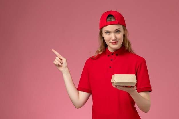 Mensajero femenino de vista frontal en uniforme rojo y capa sosteniendo un pequeño paquete de comida de entrega en la pared rosa, trabajo uniforme de la empresa de servicios de entrega