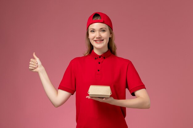 Mensajero femenino de vista frontal en uniforme rojo y capa sosteniendo un pequeño paquete de comida de entrega en la pared rosa, trabajo uniforme de la empresa de servicios de entrega