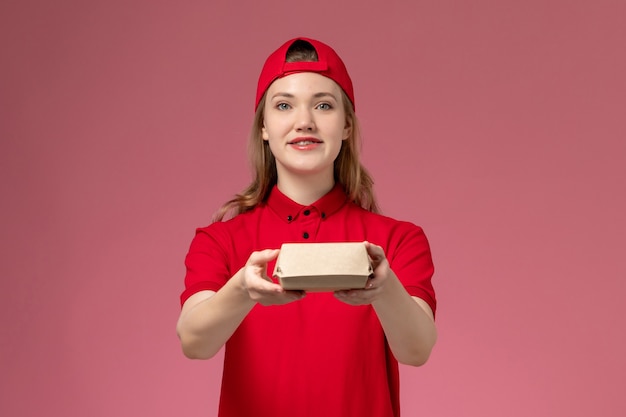 Mensajero femenino de vista frontal con uniforme rojo y capa sosteniendo un pequeño paquete de comida de entrega en la pared rosa, uniforme de empresa de servicio de trabajo de entrega