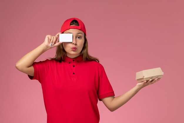 Mensajero femenino de vista frontal con uniforme rojo y capa sosteniendo un pequeño paquete de comida de entrega y una tarjeta en la pared de color rosa claro, trabajo de servicio de entrega uniforme del trabajador