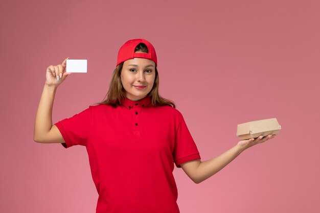 Mensajero femenino de vista frontal en uniforme rojo y capa sosteniendo un pequeño paquete de comida de entrega y una tarjeta en la pared rosa claro, trabajo de servicio de entrega uniforme