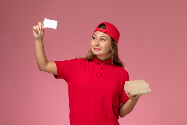 Mensajero femenino de vista frontal en uniforme rojo y capa sosteniendo un pequeño paquete de comida de entrega y una tarjeta en la pared rosa, trabajo de trabajo de servicio de entrega uniforme