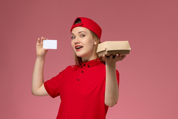 Mensajero femenino de vista frontal con uniforme rojo y capa sosteniendo un pequeño paquete de comida de entrega con tarjeta de plástico blanca en la pared de color rosa claro, servicio de entrega uniforme de trabajo