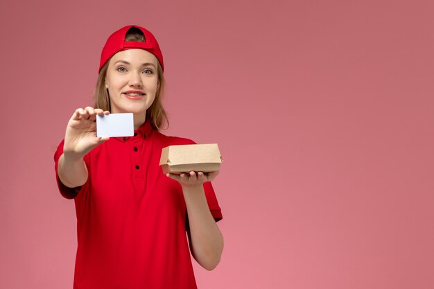 Mensajero femenino de vista frontal en uniforme rojo y capa sosteniendo un pequeño paquete de comida de entrega con tarjeta de plástico blanca sonriendo en la pared rosa, servicio de entrega uniforme de trabajo