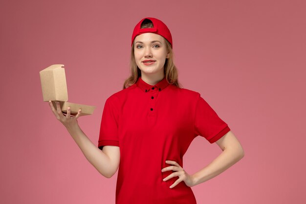 Mensajero femenino de vista frontal en uniforme rojo y capa sosteniendo un pequeño paquete de comida de entrega vacío en la pared rosa, trabajo uniforme de la empresa de servicios de entrega