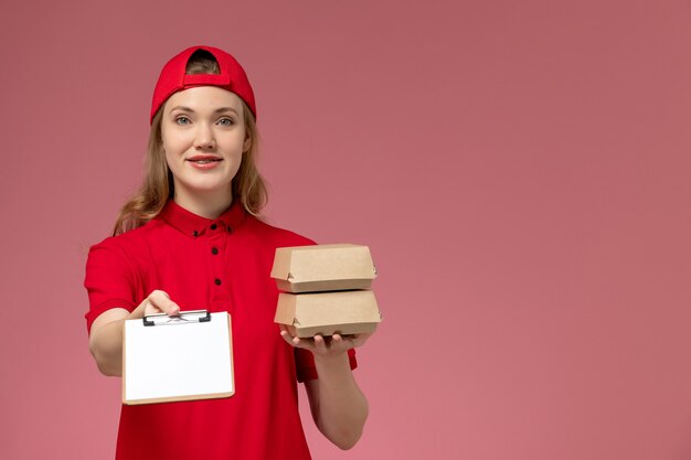 Mensajero femenino de vista frontal en uniforme rojo y capa sosteniendo pequeños paquetes de comida de entrega y bloc de notas en la pared rosa claro, servicio de entrega uniforme del trabajador