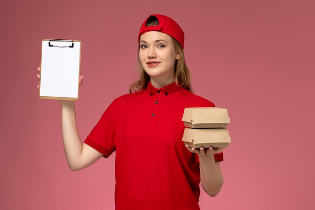 Mensajero femenino de vista frontal con uniforme rojo y capa sosteniendo pequeños paquetes de comida de entrega y bloc de notas en la pared rosa claro, servicio de entrega uniforme