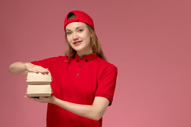 Mensajero femenino de vista frontal en uniforme rojo y capa sosteniendo pequeños paquetes de comida de entrega con una leve sonrisa en la pared de color rosa claro, servicio de entrega uniforme