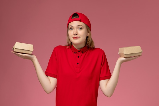 Mensajero femenino de vista frontal en uniforme rojo y capa sosteniendo pequeños paquetes de comida de entrega en la pared rosa, trabajo uniforme de la empresa de servicio de entrega del trabajador