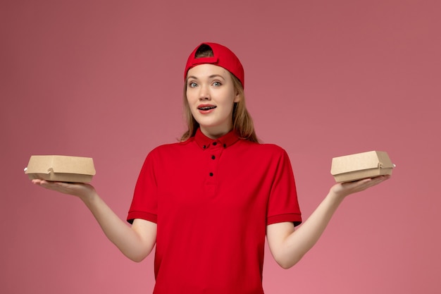 Mensajero femenino de vista frontal en uniforme rojo y capa sosteniendo pequeños paquetes de comida de entrega en la pared rosa, uniforme de trabajo de la empresa de servicio de entrega de trabajadores