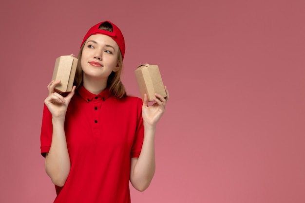 Mensajero femenino de vista frontal en uniforme rojo y capa sosteniendo pequeños paquetes de comida de entrega pensando en la pared rosa, uniforme de la empresa de servicio de entrega de trabajadores