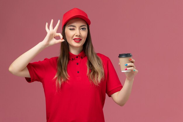 Mensajero femenino de vista frontal en uniforme rojo y capa sosteniendo la taza de café de entrega posando guiñando un ojo en el trabajador de trabajo uniforme de entrega de servicio de fondo rosa