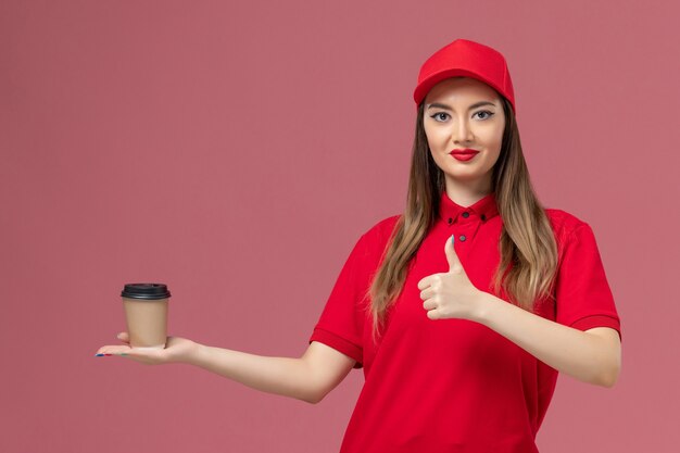 Mensajero femenino de vista frontal en uniforme rojo y capa sosteniendo la taza de café de entrega sonriendo sobre el fondo rosa trabajador de trabajo uniforme de entrega de servicio