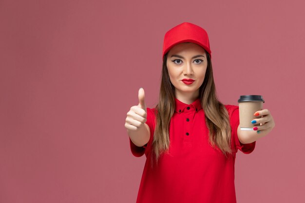 Mensajero femenino de vista frontal en uniforme rojo y capa sosteniendo la taza de café de entrega en el trabajador de trabajo uniforme de entrega de servicio de fondo rosa