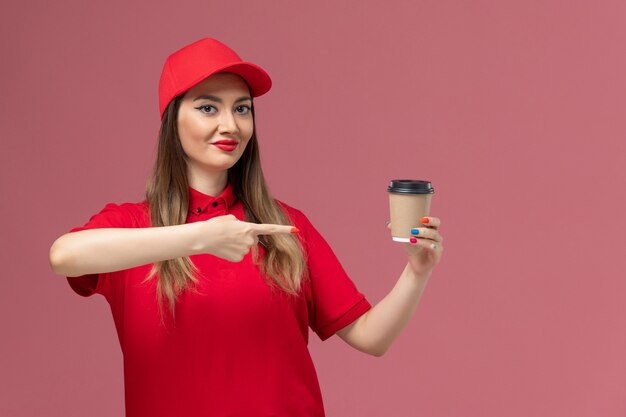 Mensajero femenino de vista frontal en uniforme rojo y capa sosteniendo la taza de café de entrega en el trabajador de trabajo uniforme de entrega de servicio de piso rosa