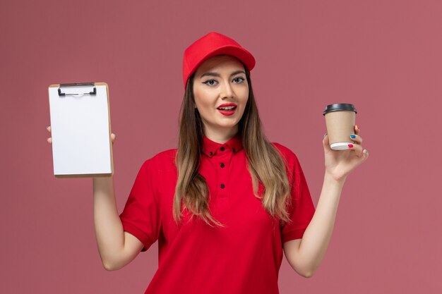 Mensajero femenino de vista frontal en uniforme rojo y capa con taza de café de entrega con bloc de notas sobre fondo rosa uniforme de trabajador de entrega de servicio