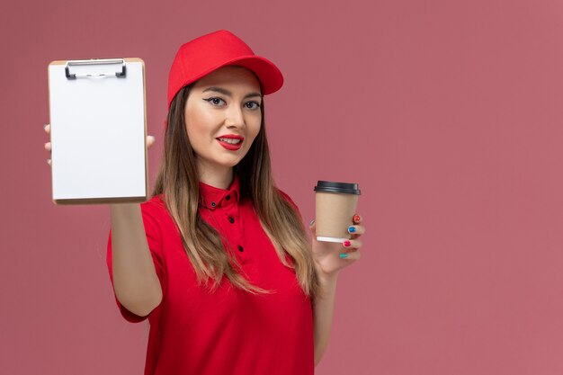 Mensajero femenino de vista frontal en uniforme rojo y capa con taza de café de entrega con bloc de notas en el uniforme de entrega de trabajo de servicio de fondo rosa claro