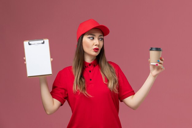 Mensajero femenino de vista frontal en uniforme rojo y capa con taza de café de entrega con bloc de notas en el uniforme de entrega de trabajo de servicio de fondo rosa