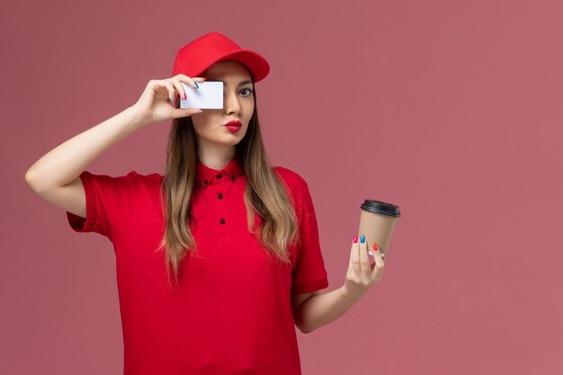 Mensajero femenino de vista frontal en uniforme rojo y capa con taza de café de entrega con tarjeta blanca en el uniforme de entrega de servicio de escritorio rosa