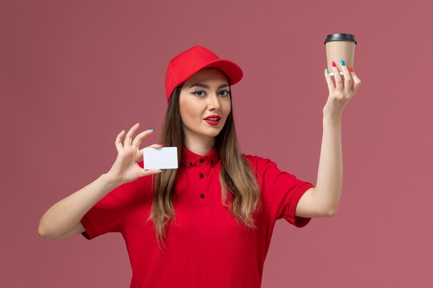 Mensajero femenino de vista frontal en uniforme rojo y capa con taza de café de entrega con tarjeta blanca en el uniforme de entrega de servicio de fondo rosa