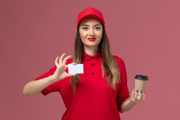 Mensajero femenino de vista frontal en uniforme rojo y capa con taza de café de entrega con tarjeta blanca en el uniforme de entrega de servicio de fondo rosa