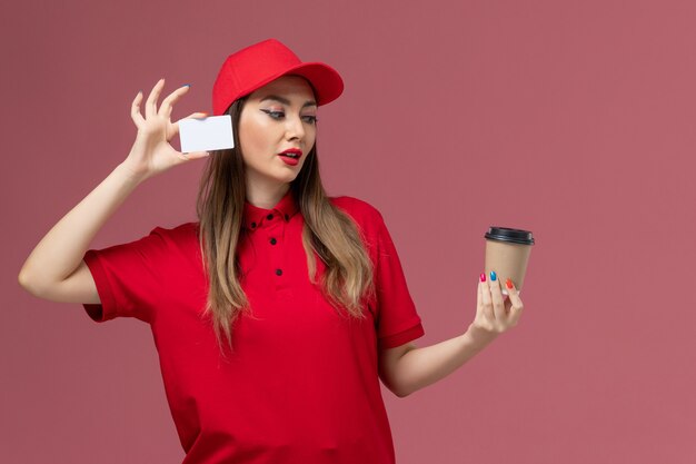 Mensajero femenino de vista frontal en uniforme rojo y capa con taza de café de entrega con tarjeta blanca en el uniforme de entrega de trabajo de servicio de fondo rosa