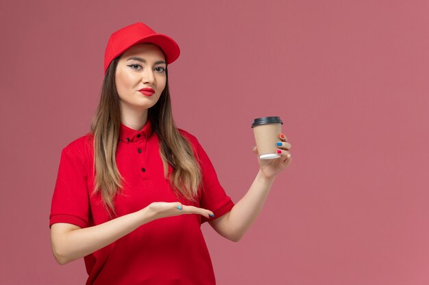 Mensajero femenino de vista frontal en uniforme rojo y capa con taza de café de entrega en el trabajador de trabajo uniforme de entrega de servicio de fondo rosa claro