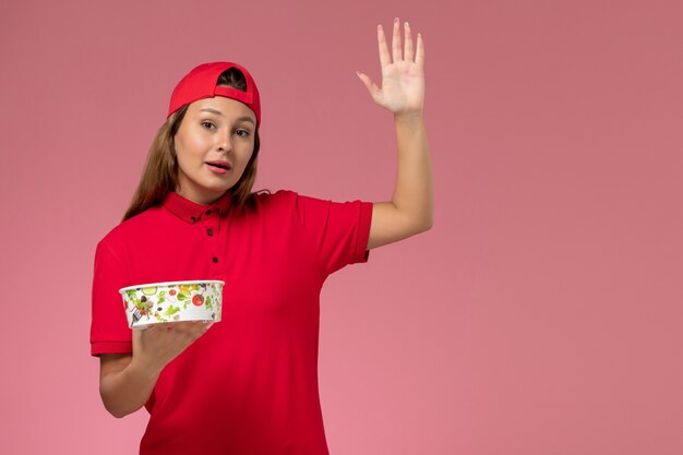 Mensajero femenino de vista frontal en uniforme rojo y capa con tazón de entrega en la pared rosa, trabajo de trabajador de servicio de entrega uniforme