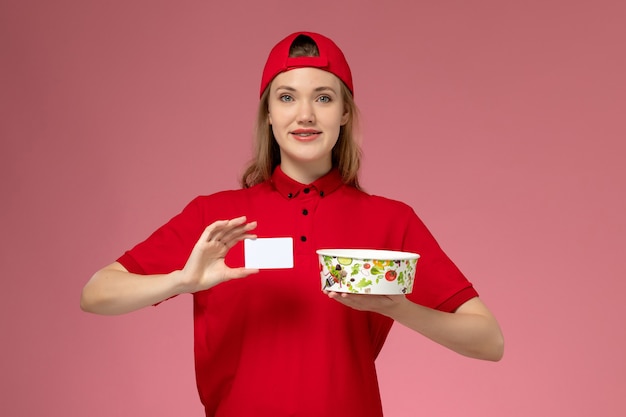 Mensajero femenino de vista frontal en uniforme rojo y capa con tazón de entrega con tarjeta en la pared rosa claro, entrega uniforme de servicio de trabajador