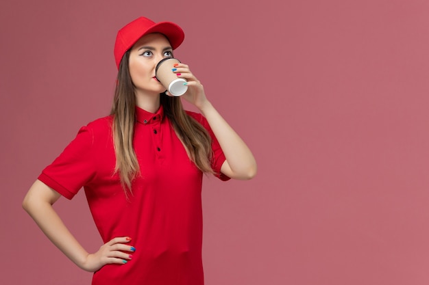 Mensajero femenino de vista frontal en uniforme rojo y capa tomando café en el trabajador de trabajo uniforme de entrega de servicio de fondo rosa