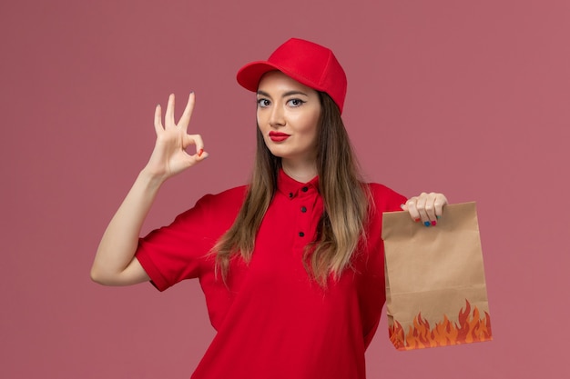 Mensajero femenino de vista frontal en uniforme rojo con paquete de comida de papel en la empresa uniforme de entrega de trabajo de servicio de fondo rosa claro