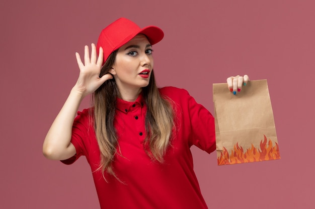 Foto gratuita mensajero femenino de vista frontal en uniforme rojo con paquete de comida de papel tratando de escuchar en el trabajo de empresa uniforme de entrega de servicio de trabajador de fondo rosa