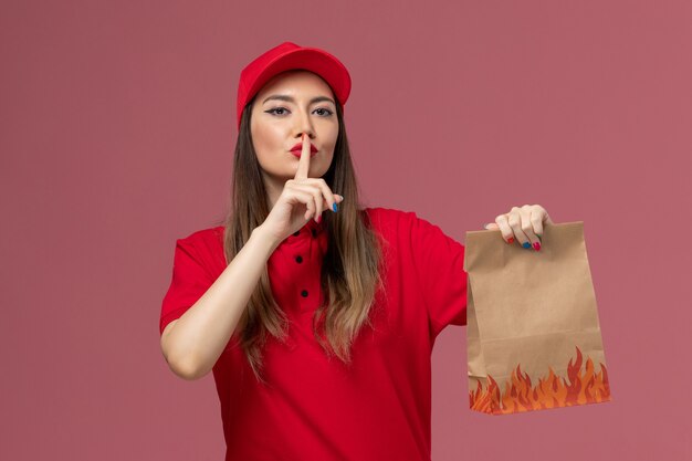 Mensajero femenino de la vista frontal en uniforme rojo que sostiene el paquete de comida de papel que muestra el signo del silencio en la compañía uniforme de la entrega del trabajo del servicio del fondo rosado