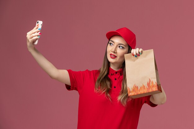 Mensajero femenino de la vista frontal en uniforme rojo que sostiene el teléfono y el paquete de alimentos que toman la foto en el trabajador de trabajo uniforme de entrega de servicio de fondo rosa