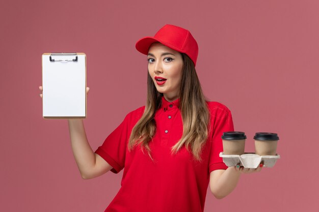 Mensajero femenino de vista frontal en uniforme rojo sosteniendo tazas de café de entrega y bloc de notas en uniforme de trabajador de trabajo de entrega de servicio de fondo rosa claro